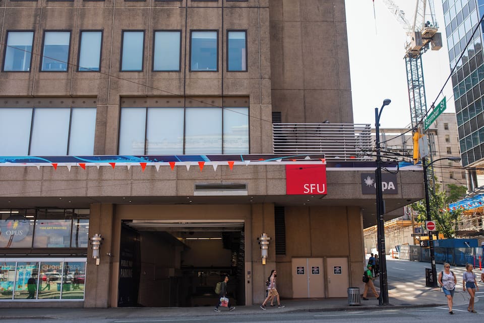 Un edificio con un letrero 'SFU', una calle con peatones y una grúa de construcción al fondo.
