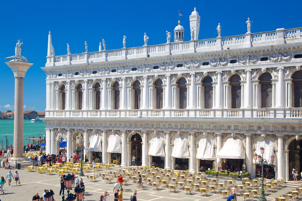 A grand white building with statues and columns, a large open plaza with yellow tables, and a clear blue sky.