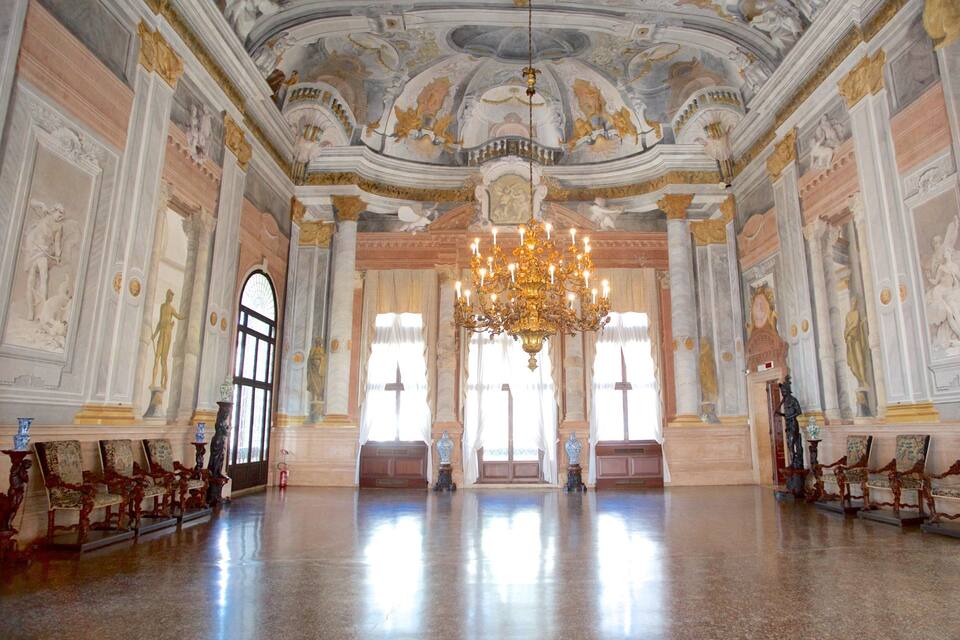 An ornate hall with a large chandelier, classical statues, and detailed frescoes on the ceiling.