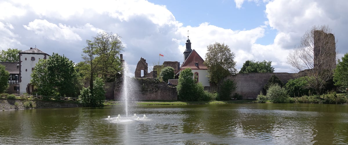 A historic castle with a fountain, surrounded by lush greenery and a calm lake.