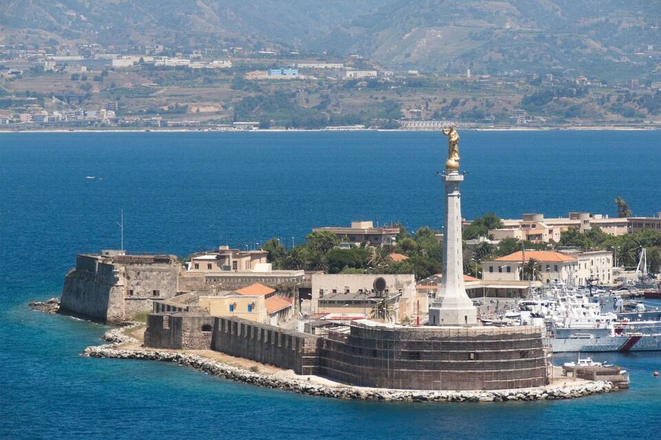 A coastal fortification with a prominent tower, surrounded by buildings and boats, with a mountainous backdrop.