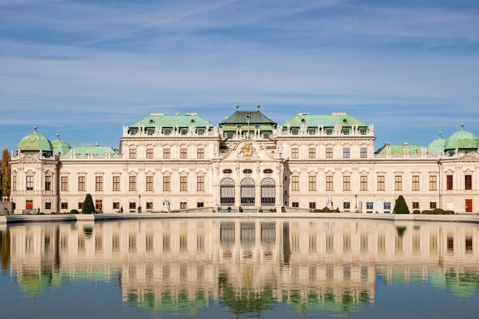 A large, ornate building with green domes and a central entrance, reflecting in a calm body of water.