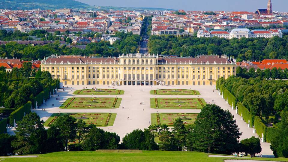 Um palácio imponente com jardins simétricos e paisagem urbana ao fundo.