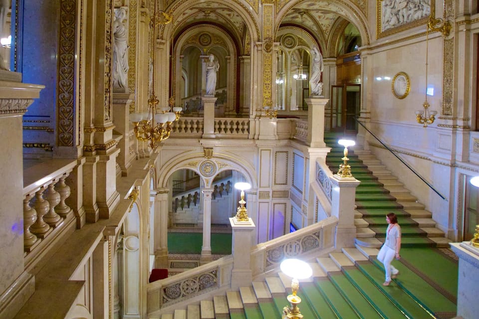 An ornate staircase with green carpet, gold chandeliers, and statues.