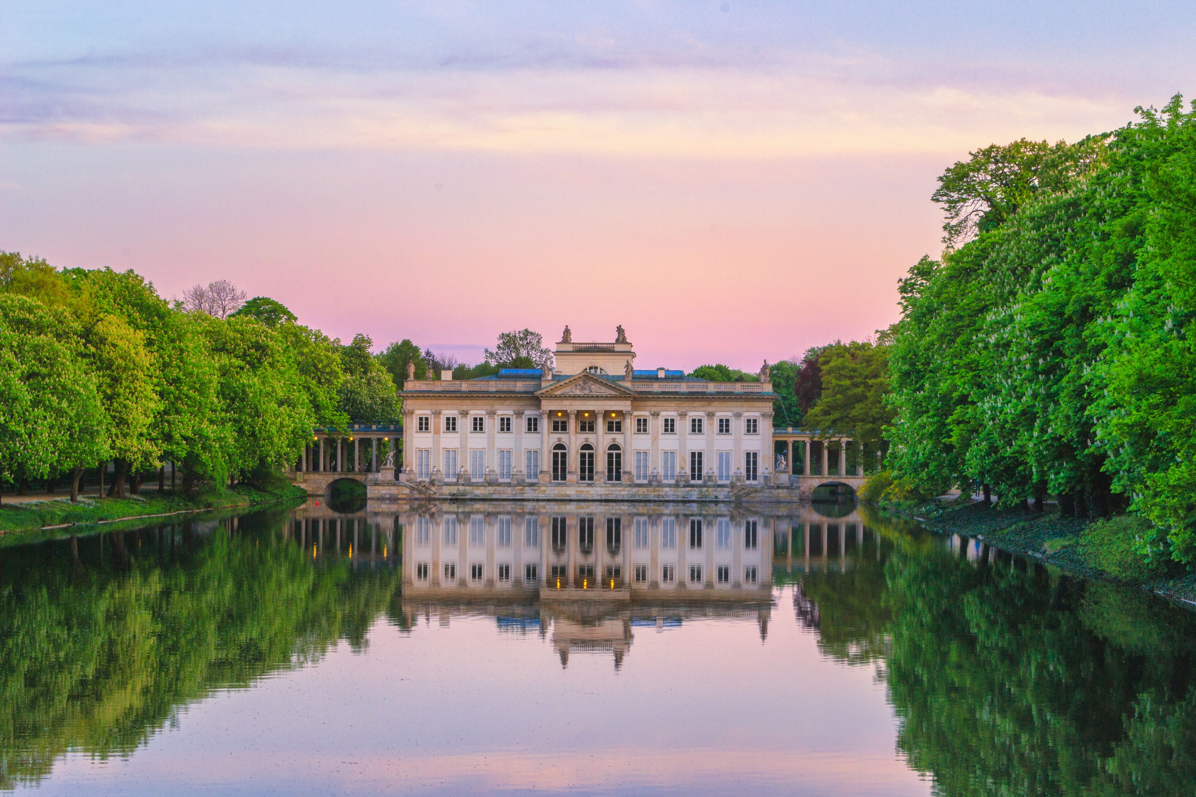 A grand building with a reflection in a calm body of water, surrounded by lush greenery.
