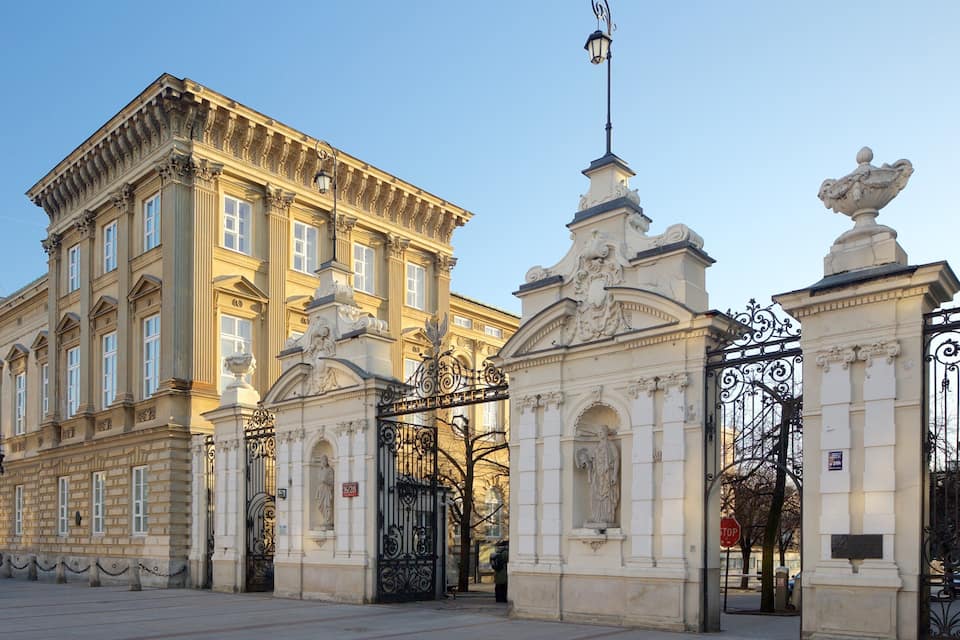 A grand classical building with ornate columns and statues, a large gate with intricate ironwork, and a clear blue sky.