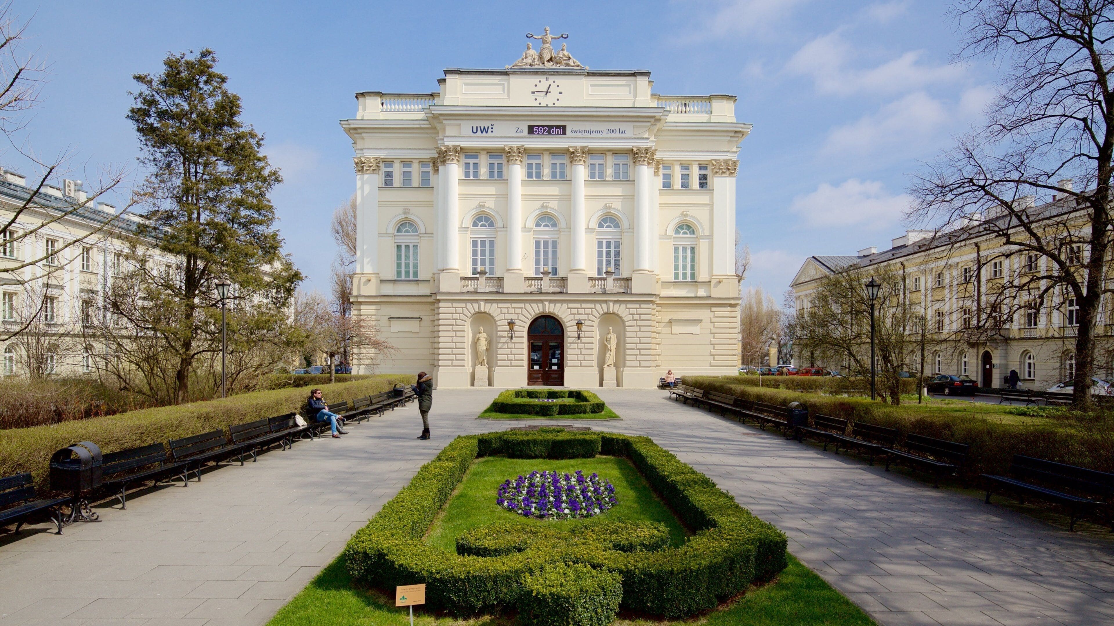 A grand building with a central entrance, flanked by symmetrical gardens and benches.