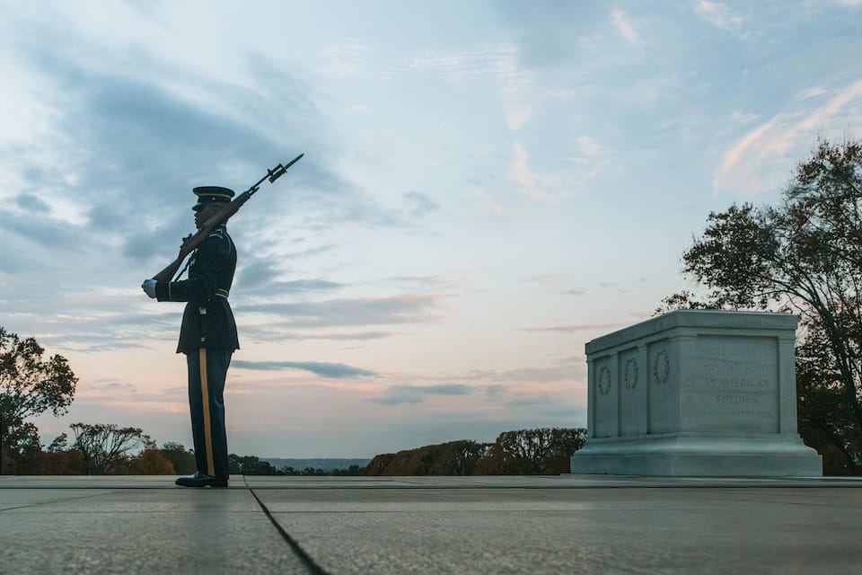 A soldier in uniform standing at attention with a rifle, a monument with inscriptions, and a clear sky with clouds.