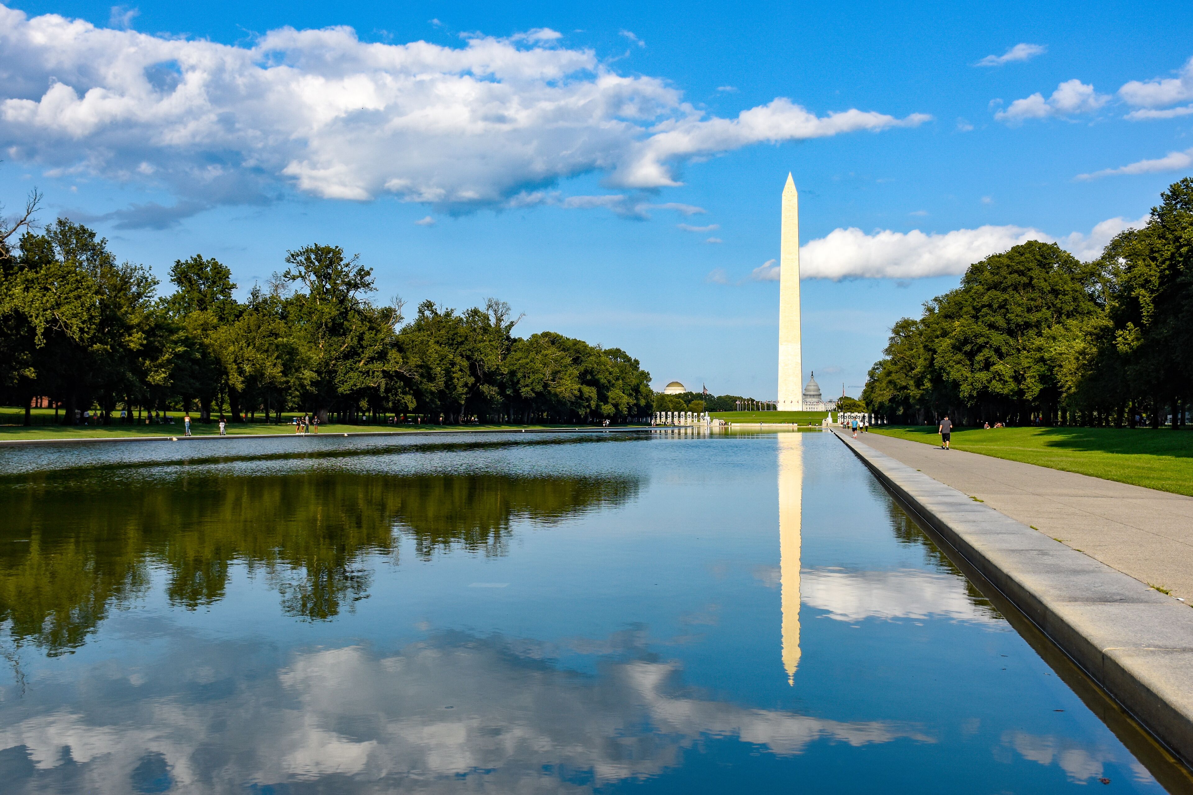 El monumento a Washington reflejado en aguas tranquilas, rodeado de frondosos árboles verdes y un cielo azul despejado.