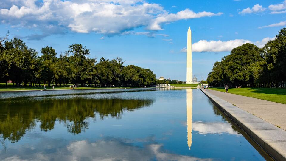 El monumento a Washington reflejado en aguas tranquilas, rodeado de frondosos árboles verdes y un cielo azul despejado.