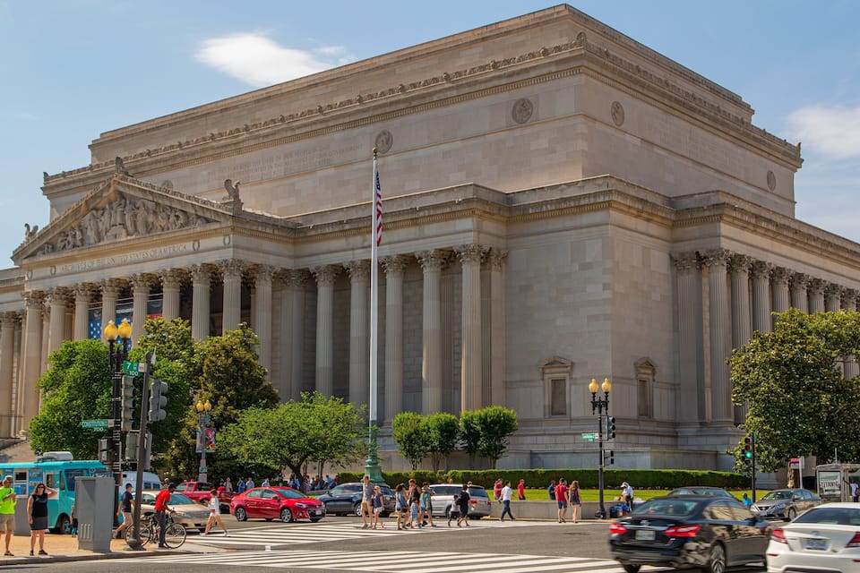 A large neoclassical building with columns and inscriptions, located at a busy intersection with pedestrians and vehicles.