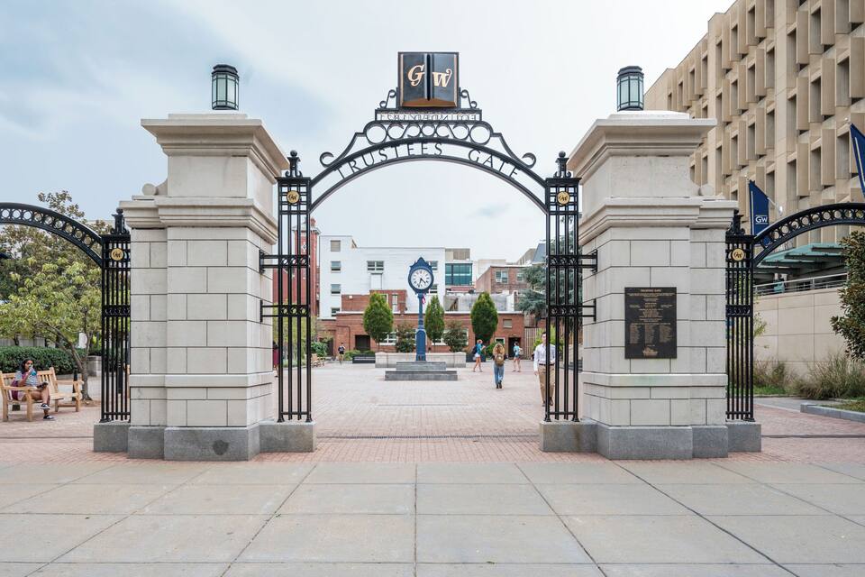 A historic gate with a clock and plaques, surrounded by a paved area and buildings.