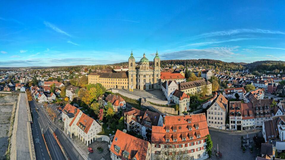 A panoramic view of a historic town with a prominent church featuring twin spires and a large dome, surrounded by traditional European architecture and a clear blue sky.
