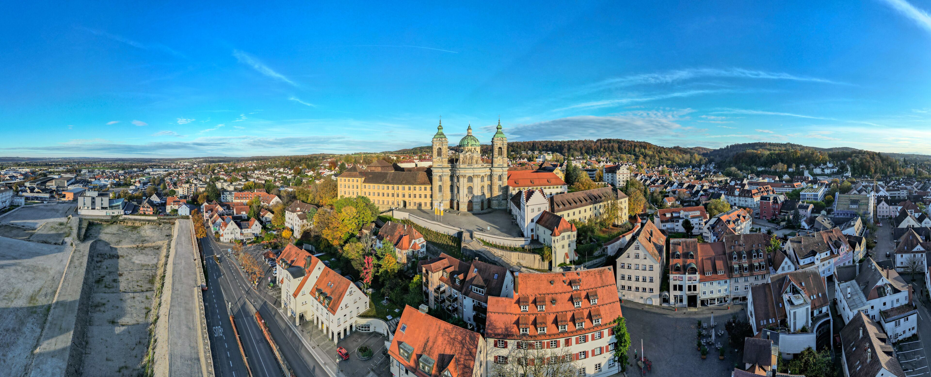 A panoramic view of a historic town with a prominent church featuring twin spires and a large dome, surrounded by traditional European architecture and a clear blue sky.
