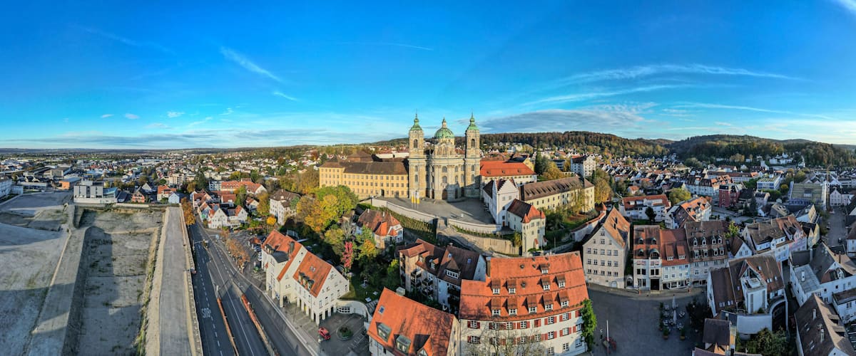 A panoramic view of a historic town with a prominent church featuring twin spires and a large dome, surrounded by traditional European architecture and a clear blue sky.