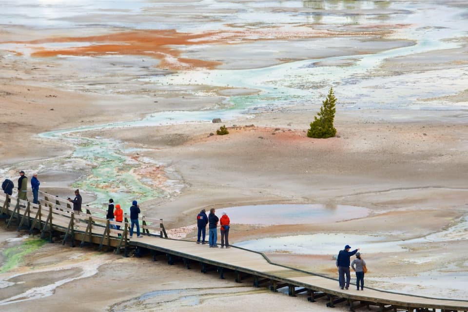 A wooden boardwalk with people observing a colorful geothermal area.