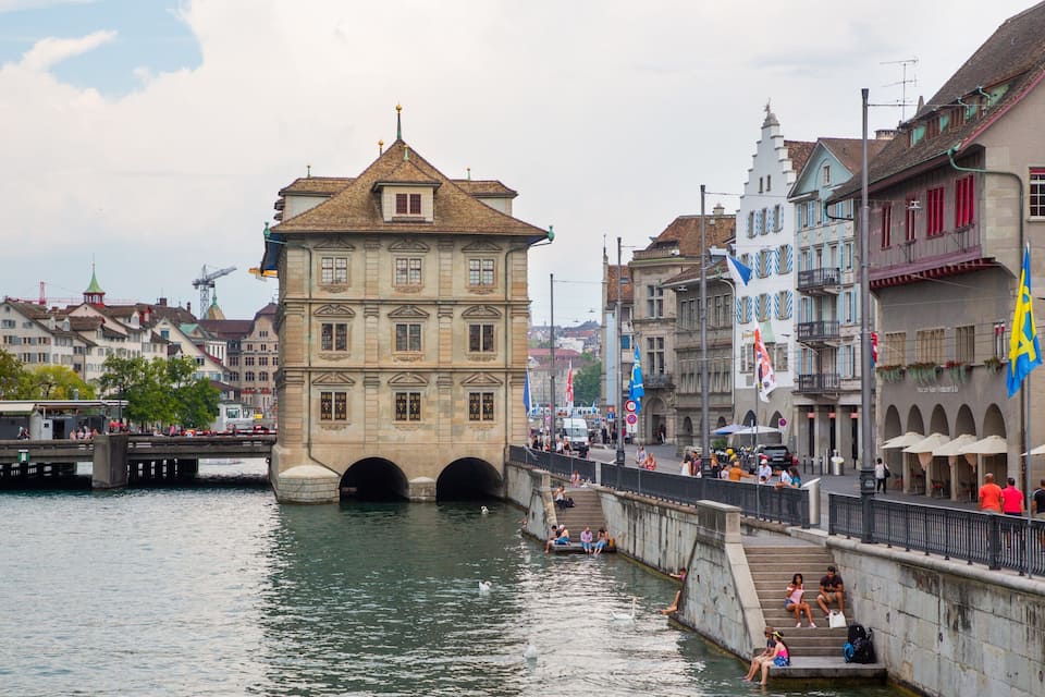 Eine Uferpromenade mit historischen Gebäuden, einer Brücke und einem Restaurant namens "Haus zum Rüden".