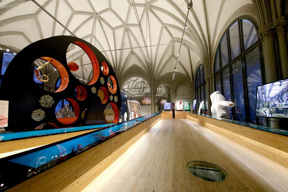 A museum interior with a large circular sculpture, wooden floors, and arched ceilings.