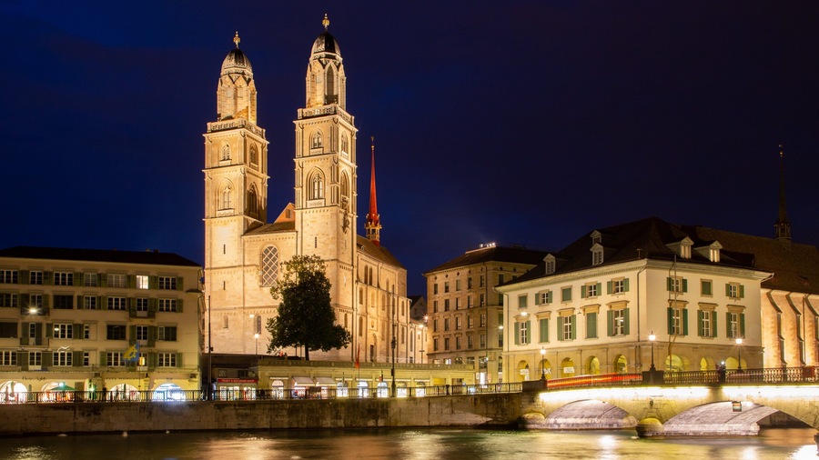 A historic cathedral with twin towers and a red spire, surrounded by classical architecture and a river.