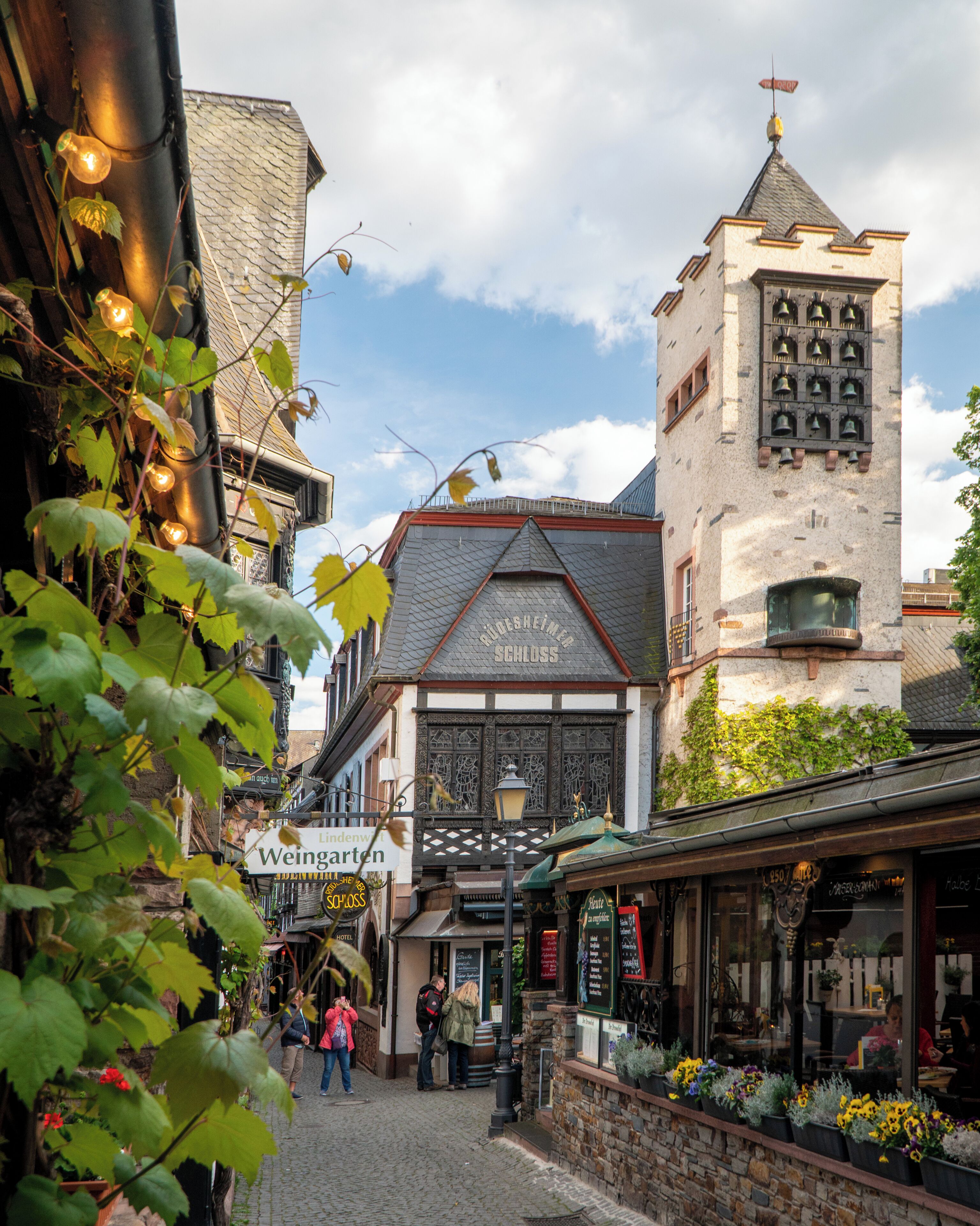A cobblestone street with historic buildings, a tower with bells, and a flower-adorned shop.