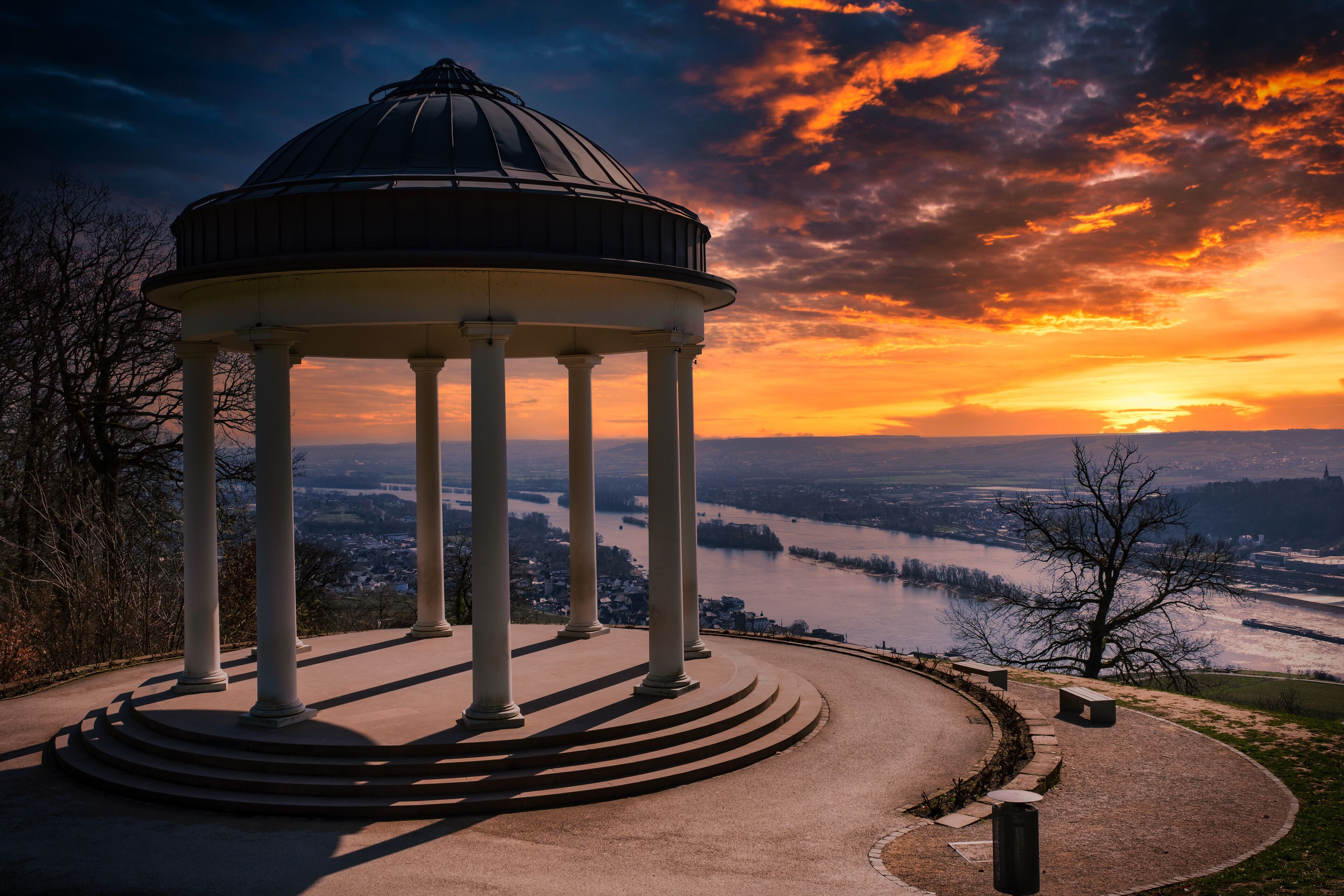 A gazebo with a dome roof, surrounded by a circular walkway, overlooking a river and a valley with buildings.