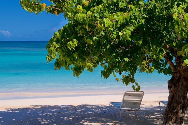 Une plage avec une eau turquoise et limpide, du sable blanc et deux chaises de plage blanches placées sous un arbre.