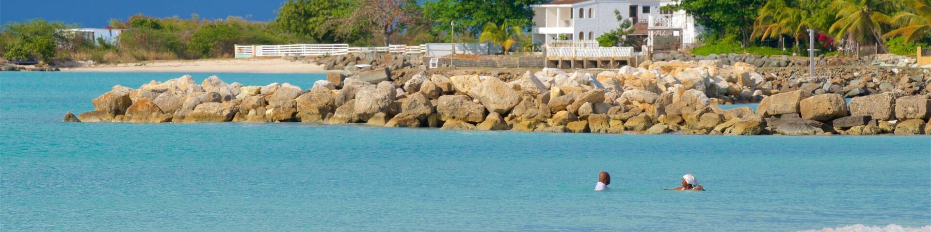 Une plage à l’eau turquoise et limpide, un rivage rocheux et, au loin, une maison blanche avec un toit rouge.