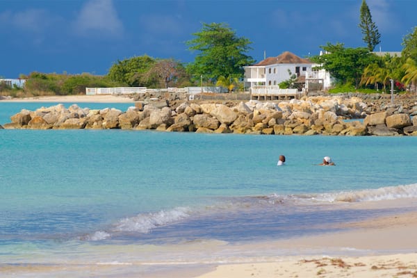 Ein Strand mit klarem türkisblauem Wasser, einer felsigen Küstenlinie und einem weißen Haus mit rotem Dach im Hintergrund.