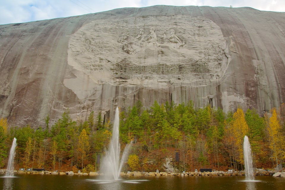 Uma grande formação rochosa com relevo esculpido, um lago com fontes e árvores em tons de outono.