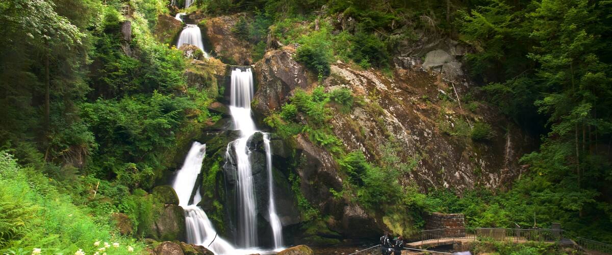 A multi-tiered waterfall surrounded by lush greenery and a viewing platform with people.