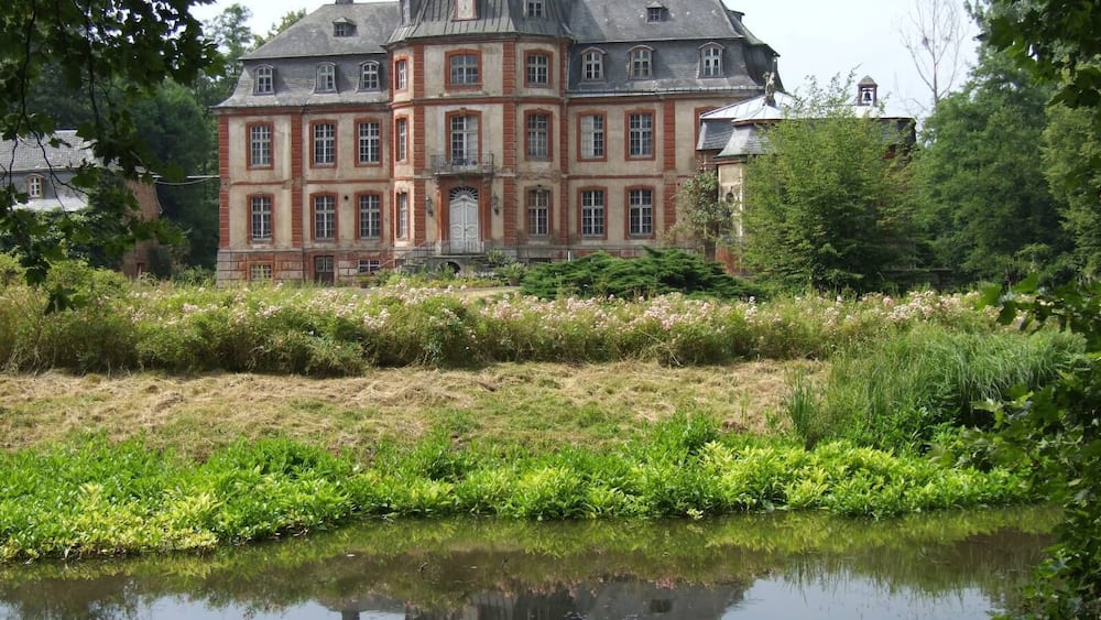 Un grand bâtiment historique à la façade symétrique, entouré d’une végétation luxuriante et bordé d’un étang paisible dans lequel se reflète son image.