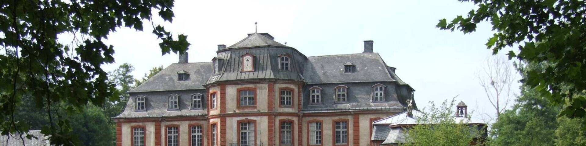 A large, historic building with a symmetrical facade, surrounded by lush greenery and a calm pond reflecting its image.