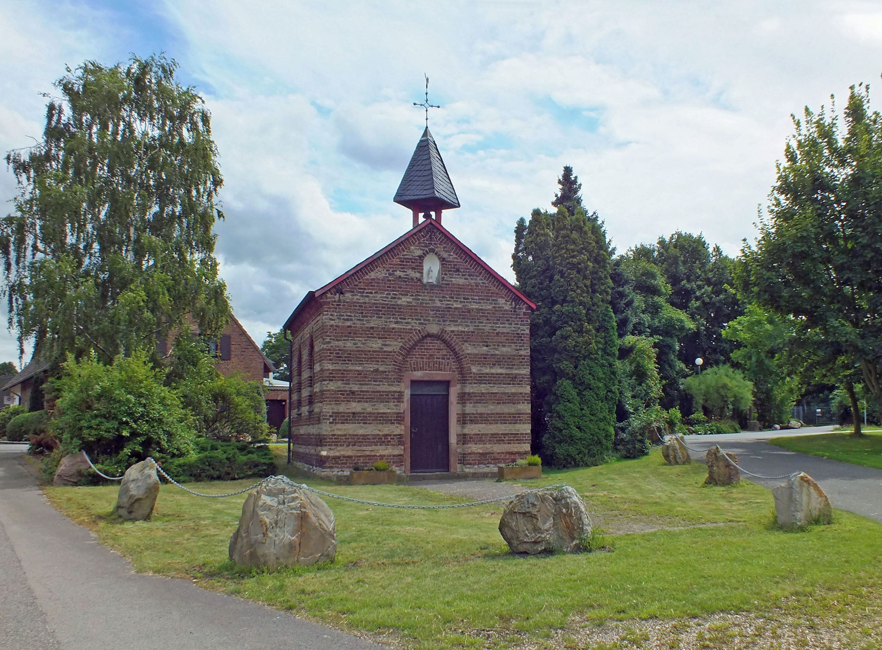A small brick church with a pointed steeple and a cross on top, surrounded by a grassy area with large rocks and trees.