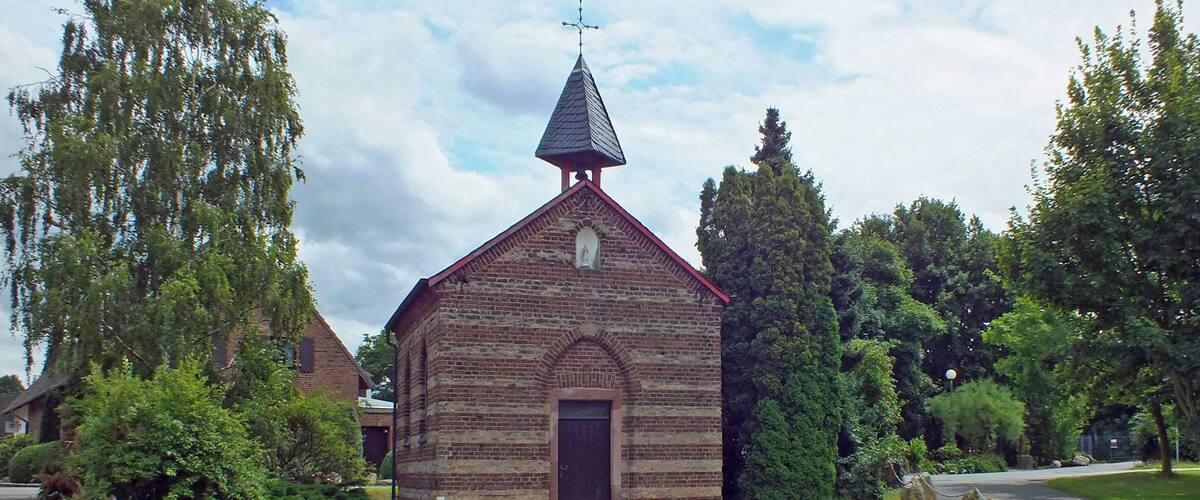A small brick church with a pointed steeple and a cross on top, surrounded by a grassy area with large rocks and trees.