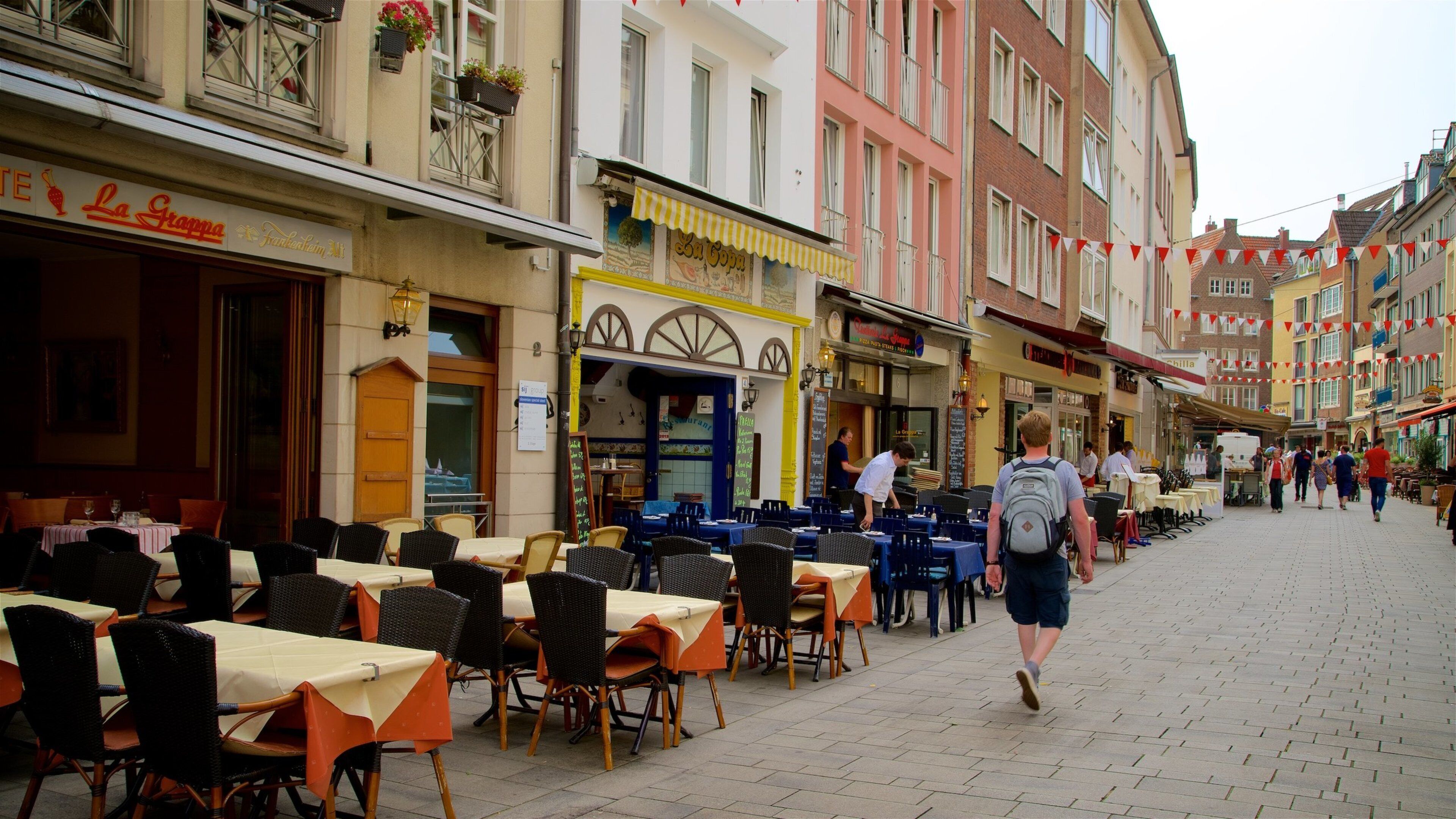 A cobblestone street lined with outdoor cafes and restaurants, a pedestrian, and colorful buildings.