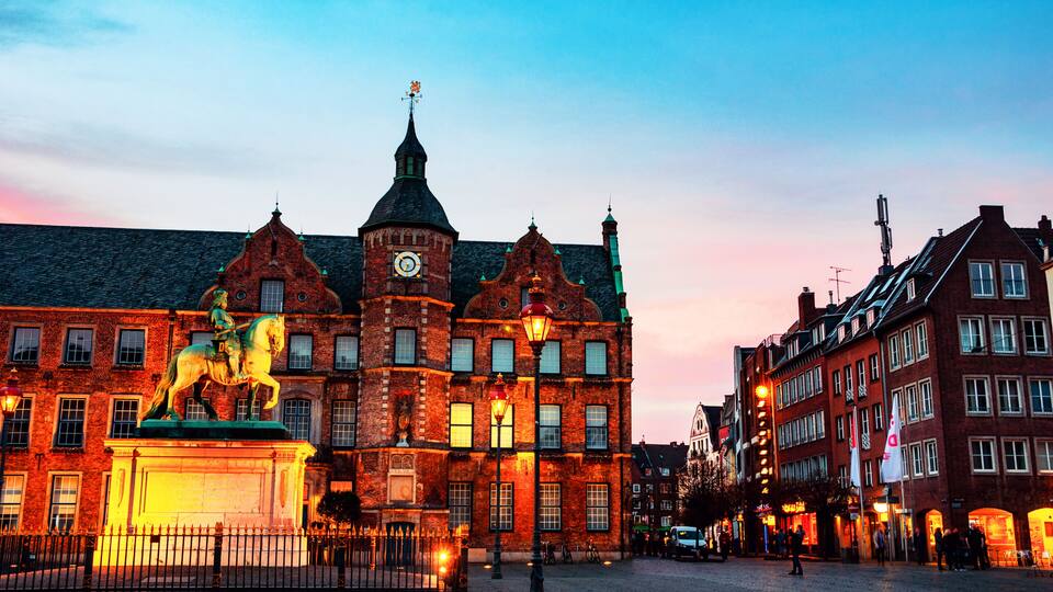 Een historisch gebouw met een klokkentoren, een standbeeld van een ruiter en een geplaveid plein.