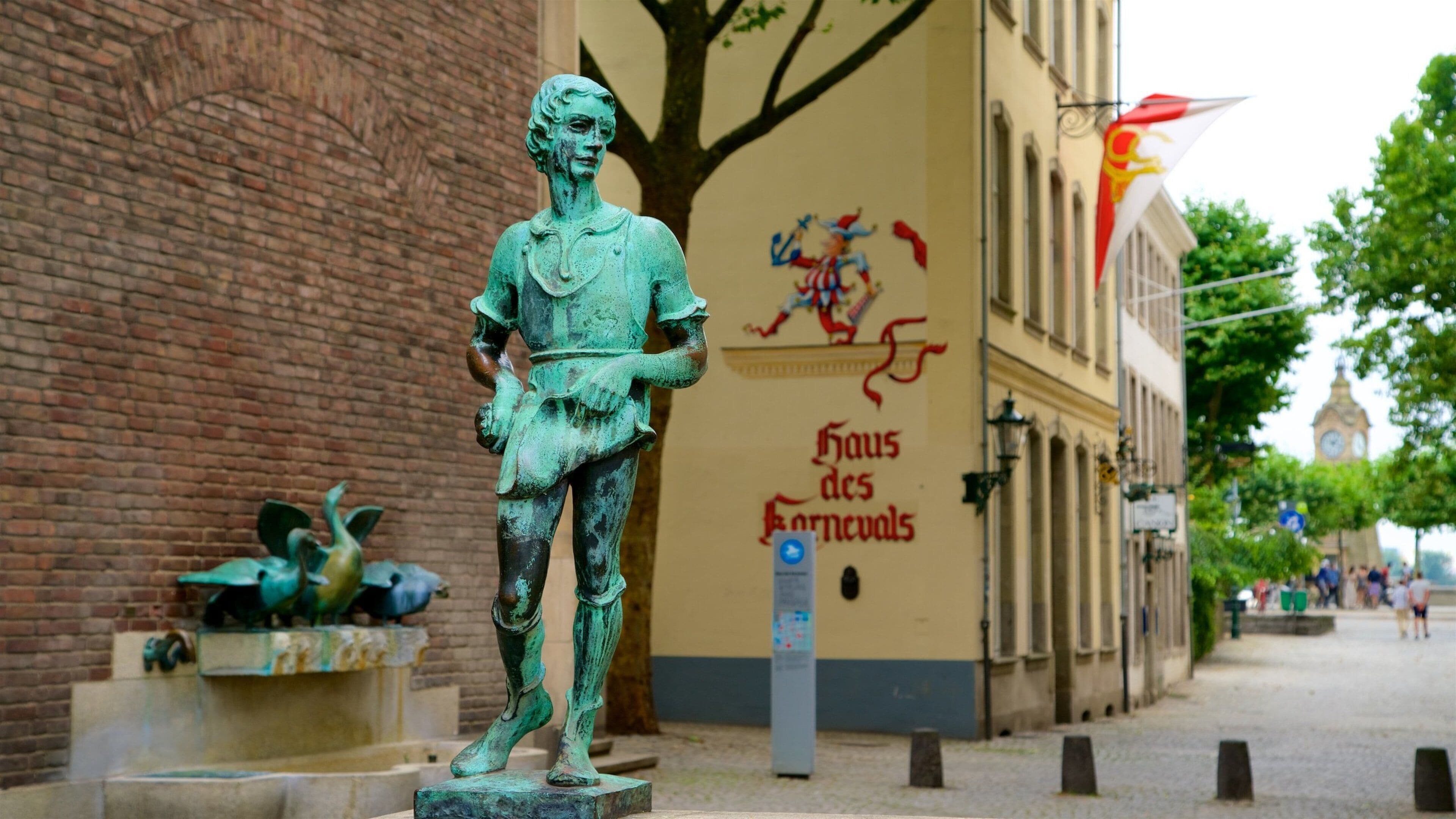 A statue of a figure in a greenish patina, a brick wall, and a building with a clock tower in the background.