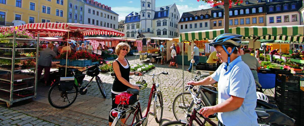 A bustling outdoor market with vendors and bicycles, surrounded by historic buildings and a prominent clock tower.