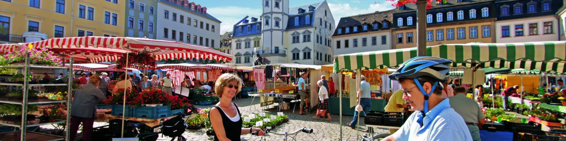 A bustling outdoor market with vendors and bicycles, surrounded by historic buildings and a prominent clock tower.