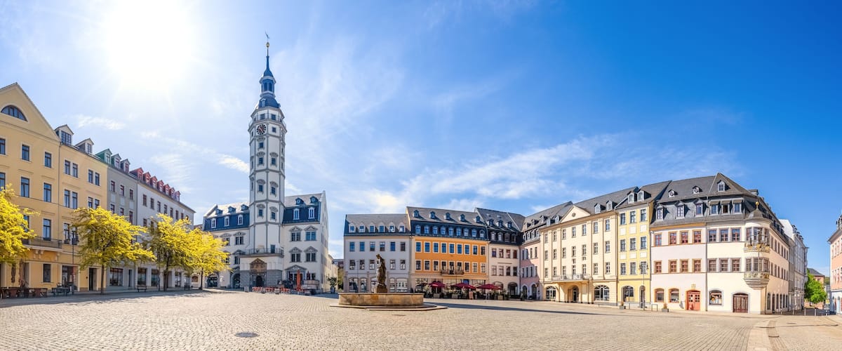Uma praça de paralelepípedos com edifícios históricos, uma torre do relógio imponente e uma estátua no centro.