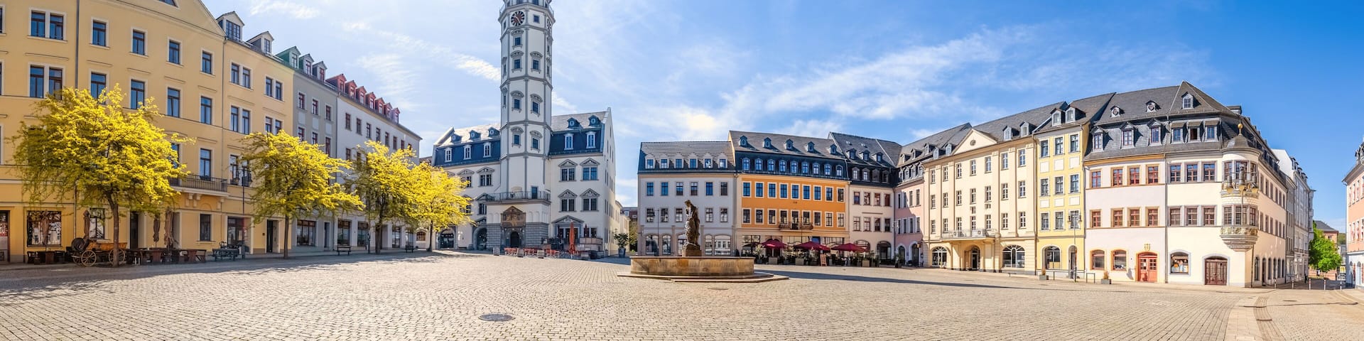 A cobblestone square with historic buildings, a prominent clock tower, and a statue in the center.