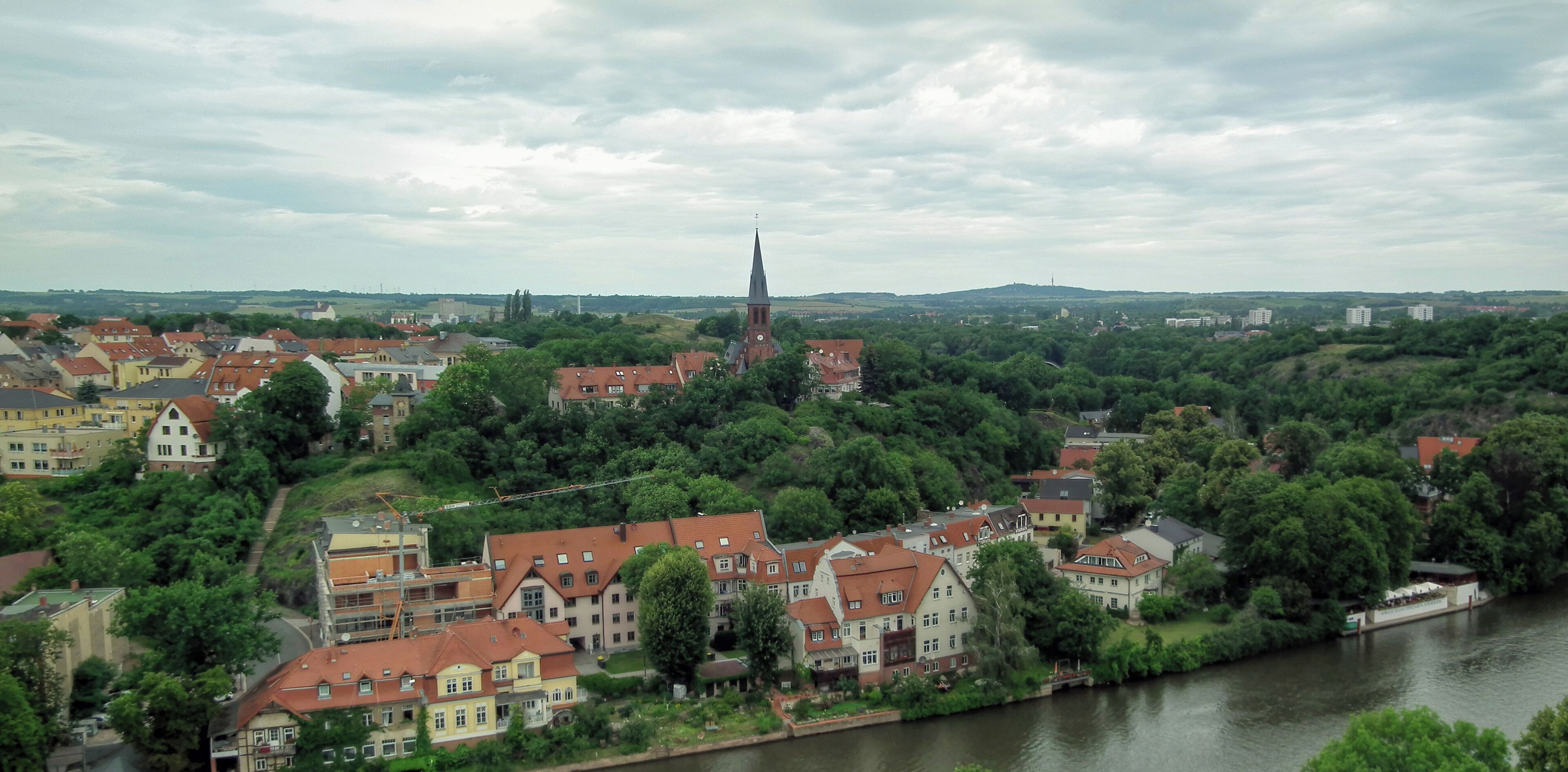A town with a prominent church steeple, a river, and surrounding greenery.