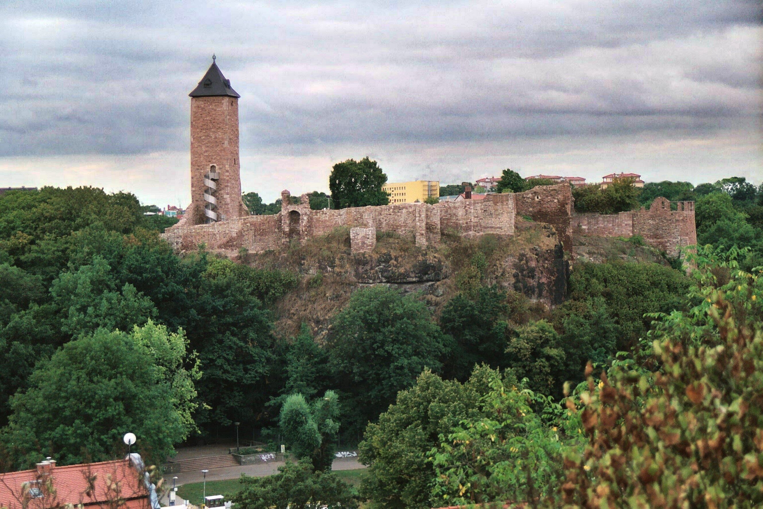 A historic stone castle with a tall tower, surrounded by lush greenery and modern buildings in the background.
