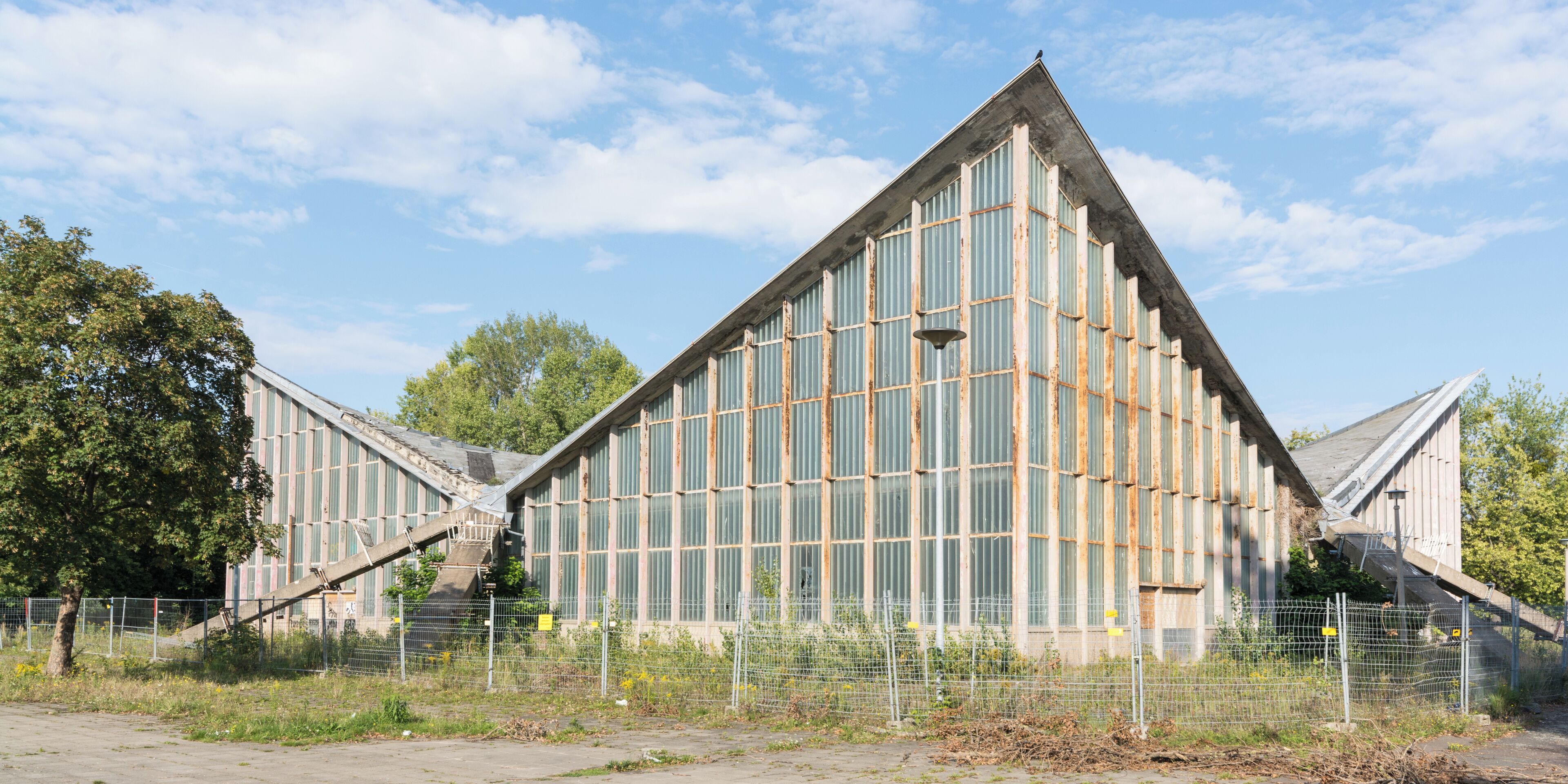 A large, triangular building with a metal exterior and large windows, surrounded by a fence and trees.