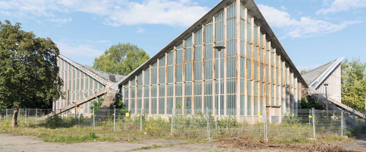 A large, triangular building with a metal exterior and large windows, surrounded by a fence and trees.