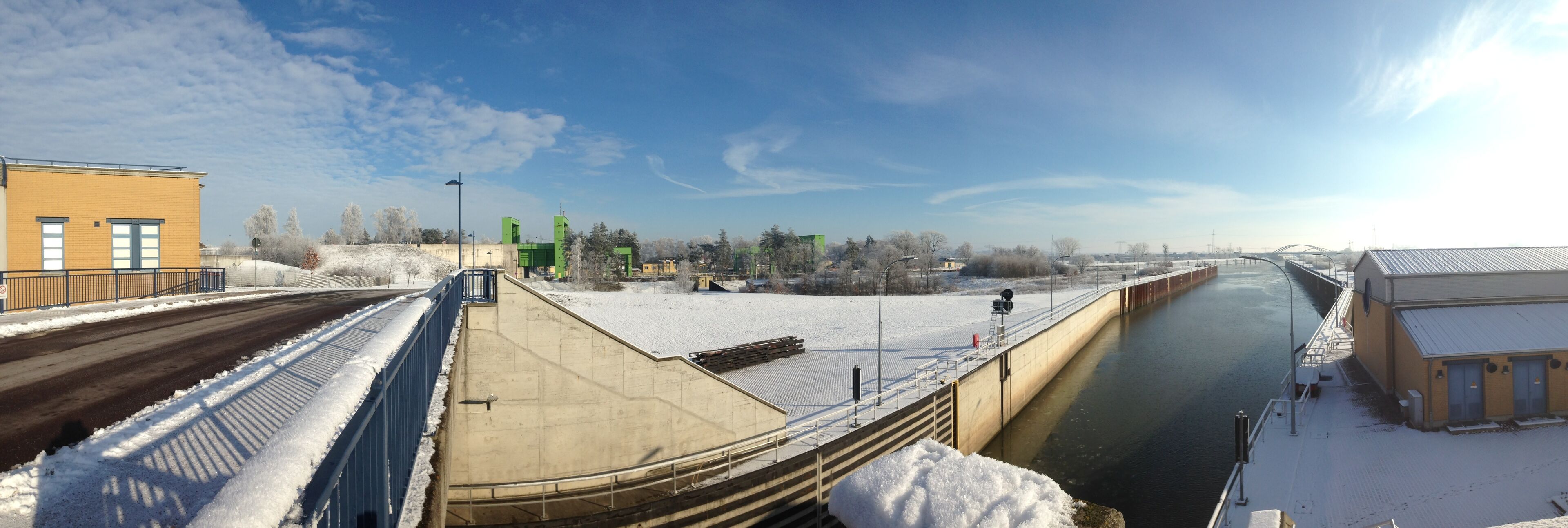 A snow-covered dam with a river flowing through it, a bridge, and some buildings.