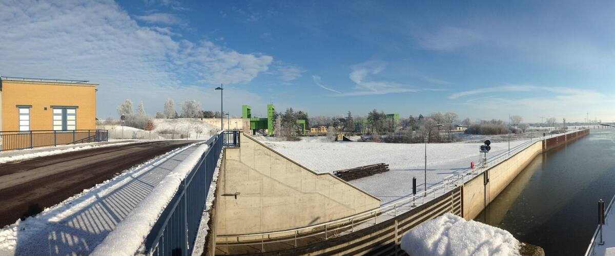 A snow-covered dam with a river flowing through it, a bridge, and some buildings.