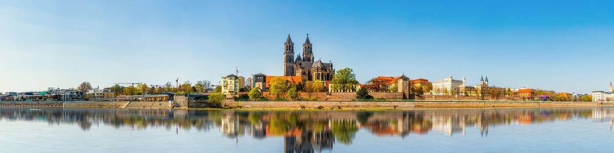 Eine historische Kathedrale mit zwei Türmen, ein ruhiger Fluss im Vordergrund und eine Mischung aus modernen und traditionellen Gebäuden entlang des Ufers.