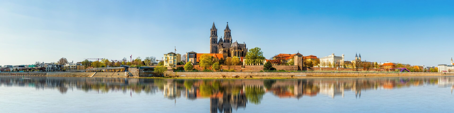 A historic cathedral with twin spires, a calm river in the foreground, and a mix of modern and traditional buildings along the riverbank.
