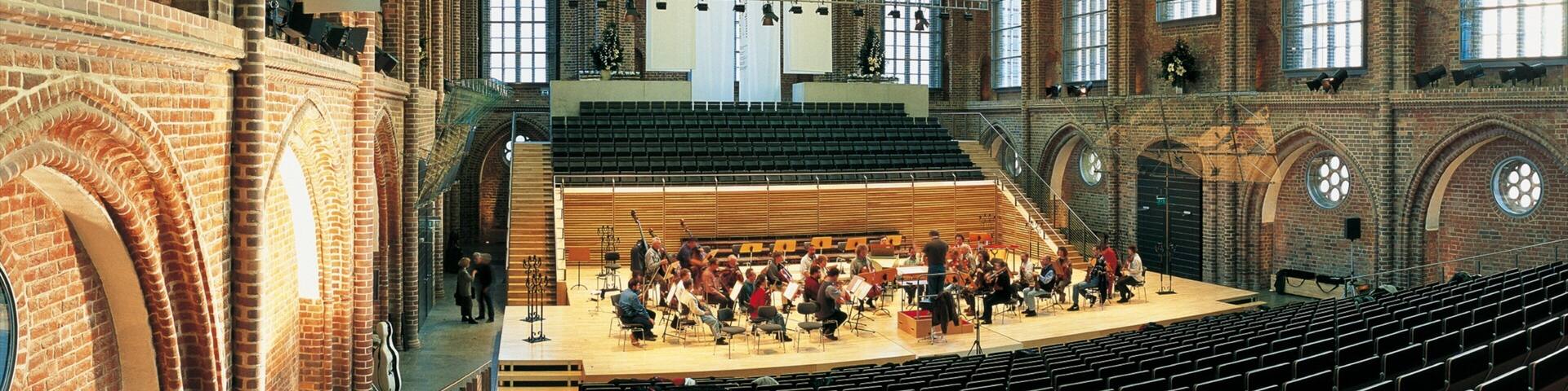 An indoor concert hall with a high arched ceiling, brick walls, and large windows.